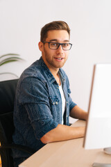 Vertical shot of confident young freelance programmer male in stylish glasses working on desktop computer sitting at desk at home office, in room with modern interior, looking at camera.