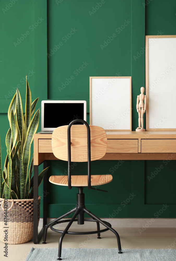 Modern workplace with laptop, mannequin and blank frames near green wall