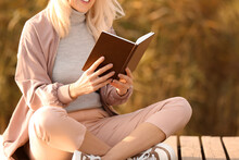 Mature Woman Reading Book Near River On Autumn Day