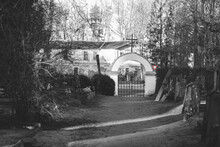 Pathway Out Of Cemetery. Cemetery Gate With Arc And Catholic Cross, Give Way Road Sign And Old Abanoned Building Outside Graveyard. Black And White With Red Color Left