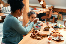 Black Little Girl Eat Breakfast With Her Mother Who Is Feeding Her At Dining Table.