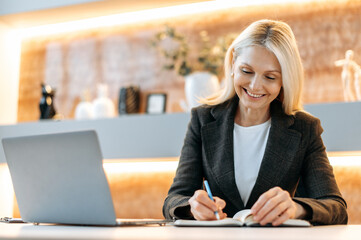 Wall Mural - Happy smart caucasian businesswoman, tom manager, ceo, sitting at a table in a modern office, using a laptop, pondering business strategies and financial opportunities, taking notes, smiling