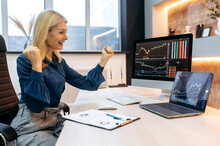 Happy Joyful Mature Blonde Business Woman, Trade Broker, Stock Trader, Sits At Table In Office, Shouts And Rejoices At Success, Good Profit, Looks At Charts On Pc And Laptop, Gestures With Fists