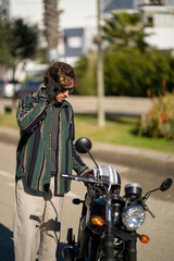 Adolescente hombre joven motero con camisa de cuadros elegante preparándose para viaje. Fondo de palmeras veraniegas. Chico elegante con gafas de sol y moto. Chico poniéndose casco de seguridad.