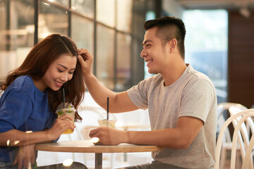 Poster - Smiling young Asian man sitting at small round table and adjusting hair to girlfriend in cafe