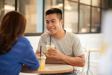 Sticker - Smiling young Asian man in tshirt sitting at table and drinking lemonade while dating with girl in cafe