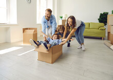 Husband And Wife With Children Who Have Just Moved Into Their Own House Are Having Fun And Rejoicing. Parents Ride Their Little Sons In Cardboard Box. Concept Of Buying Your Own Home And Moving.