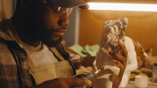 African American Craftsman In Checkered Shirt And Cap Polishing Half Of Plaster Bust While Sitting Near Illuminated Workbench And Working In Sculpting Workshop At Night
