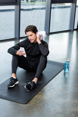 Wall Mural - Sportsman using smartphone and wiping neck with towel on fitness mat in gym