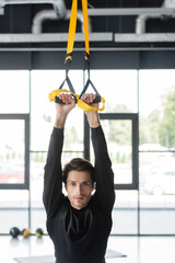 Wall Mural - Brunette sportsman training with suspension straps and looking at camera in gym