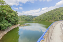 Esch-sur-Sure Dam Surrounded By Lush Trees Seen From The Country Road, Valley With Hills Covered With Green Vegetation In The Background, Sunny Day With Blue Sky White Clouds In Luxembourg