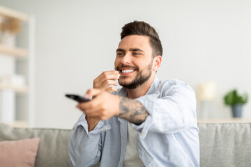 Canvas Print - Happy relaxed guy watching Tv and eating popcorn, switching channels with remote controller, resting on couch