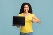 © Prostock-studio - Great website. Happy african american lady pointing at laptop with black screen, standing over blue background