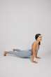 © Юля Бурмистрова - Brunette poses on isolated background. Young woman in gray tracksuit performs exercise from yoga or stretching, she stands in cobra pose and smiles, side view.