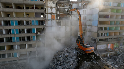 Wall Mural - Excavator for dismantling buildings, using a special claw, dismantles a multi-storey building. The sun's rays break through a thick cloud of dust in front of the ruined building