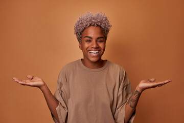 Wall Mural - Surprised joyful young African American woman looks directly at you smiling broadly spreading hands, demonstrating incomprehension and indecision standing in casual clothes on brown background