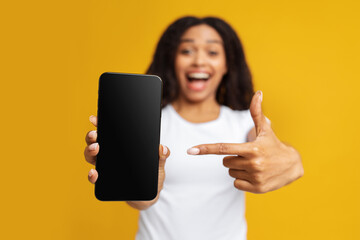 Cool mobile app. Overjoyed african american lady pointing finger at blank smartphone screen over yellow background