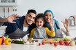 © Prostock-studio - Cheerful Islamic Family Of Three Waving At Camera While Cooking In Kitchen