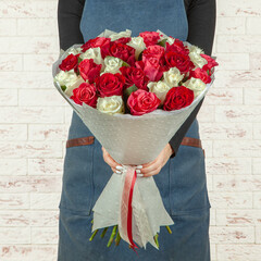 Young girl florist in a blue apron holds a fresh bouquet of red flowers wrapped in paper with a ribbon on a brick wall background