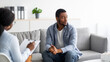 Depressed black man consulting psychologist, sitting on couch deep in thought, panorama