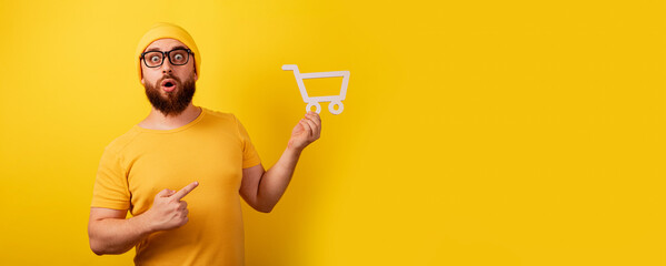 Wall Mural - shocked man pointing at shopping cart symbol in hand over yellow background, black friday and big sales concept, panoramic layout