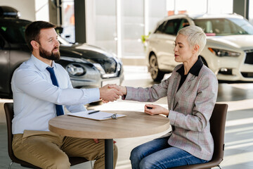 Canvas Print - Charming female customer reading car purchase document and shaking hands while sitting at table with dealer in auto salon interior.