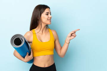 Wall Mural - Young sport woman with mat isolated on blue background pointing to the side to present a product