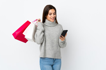 Sticker - Young caucasian woman isolated on white background holding shopping bags and writing a message with her cell phone to a friend