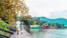Back View Of Asian Grandfather And Grandchild Wear Protective Face Mask To Prevent Coronavirus (COVID-19) Walking In A Nature Path On Wooden Bridge Along River. Together Outdoors Family Concept.