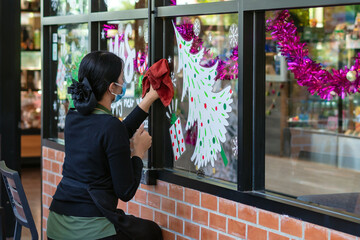 Asian woman decorates beautiful stickers on window glass in front of cafe to welcome Christmas and New Year. Prepare to decorate the storefront to be beautiful during the important holidays of year.