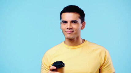 pleased young man in yellow t-shirt holding paper cup isolated on blue.