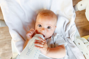 Cute little newborn girl drinking milk from bottle and looking at camera on white background. Infant baby sucking eating milk nutrition lying down on crib bed at home. Motherhood happy child concept