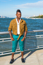 Dressing In A Yellow Woolen Jacket,  Green Pants And Black Leather Shoes,  A Young Black Guy With Mohawk Hair Is Standing By A River And Confidently Looking Forward.