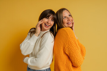 Close up portrait  of two pretty woman in cozy sweaters posing over yellow background.  Autumn and winter fashion trends.