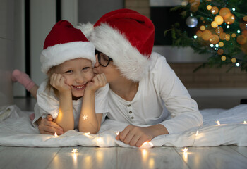 Children in santa claus hats lie under the tree. Boy kissing girl