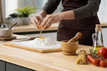 Wall Mural - Hands of young woman breaking egg on top of pile of sifted flour on wooden board while making dough