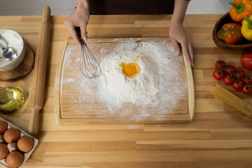 Wall Mural - Young woman holding whisk over pile of sifted flour with raw egg yolk on top while preparing dough for homemade pastry
