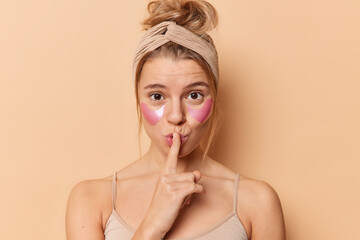 Wall Mural - Wondered woman makes hush sign presses index finger over lips applies collagen patches under eyes to reduce wrinkles or puffiness wears headband t shirt isolated over beige background. Beauty concept