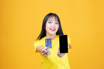 Wall Mural - Portrait Asian happy young girl smiling cheerful and showing plastic credit card while holding mobile phone isolated on yellow background