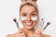 Headshot Of Smiling Young European Woman Applies Beauty Patches Under Eyes Raises Eyebrows Feels Glad Going To Apply Makeup Holds Cosmetic Brushes Wears Headband Isolated Over White Background