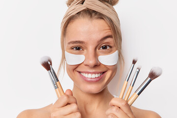 Wall Mural - Headshot of smiling young European woman applies beauty patches under eyes raises eyebrows feels glad going to apply makeup holds cosmetic brushes wears headband isolated over white background