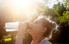 Young Blonde Sportswoman Doing Exercise Drinking Bottle Of Water Against The Background Of The Park And The Sun's Rays, The Concept Of Healthy Life And Active Loads
