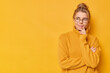 canvas print picture - Pensive dreamy young European woman keeps hand near mouth looks suspicious away wears round spectacles and jumper isolated over yellow backgroud copy space aside for your promotional content