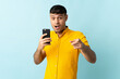 © luismolinero - Young Colombian man using mobile phone isolated om blue background surprised and pointing front
