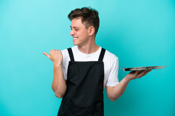 Wall Mural - Young waitress with tray isolated on blue background pointing to the side to present a product