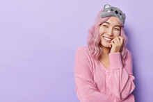 Horizontal Shot Of Dreamy Asian Woman Touches Face With Hands Has Tender Expression Smiles Joyfully Wears Casual Jumper And Sleepmask Isolated Over Puple Background Blank Copy Space. Sincere Emotions