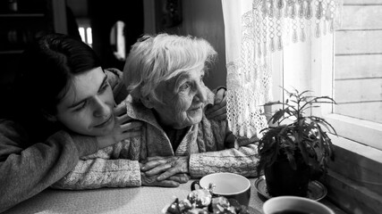 Wall Mural - An elderly woman looks at a window, with his adult granddaughter, at home. Self-isolation. Black and white photo.