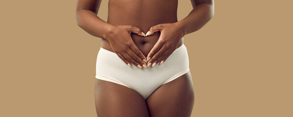 Plus size black woman in underwear holding hands on abdominal area forming heart shape around navel, isolated on solid brown color background, cropped shot. Self love, care, health, pregnancy concept