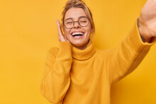 Happy Carefree Woman Smiles Gladfully Holds Camera As Taking Selfie Poses For Making Photo Pulls Wears Round Eyeglasses And Sweater Isolated Over Yellow Background Has Fun Expresses Positive Emotions