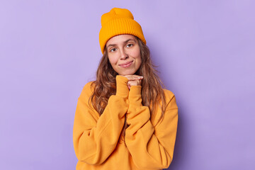 Wall Mural - Charming long haired woman looks with tender expression keeps hands under chin wears casual orange jumper and hat isolated over purple background. People face expressions and felings concept